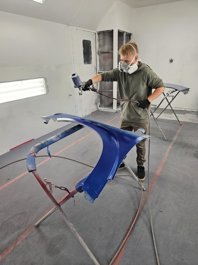 A technician in a respirator spraying a car fender metallic blue inside the spray booth