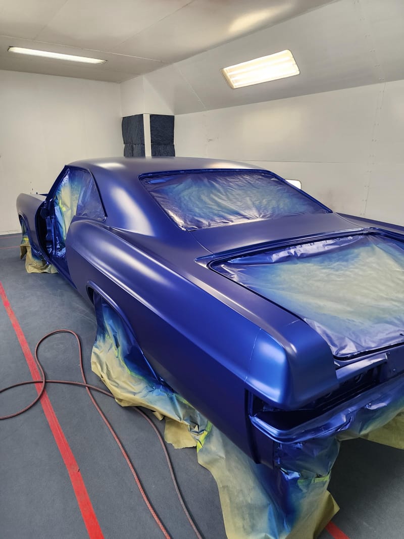 A classic two-door car freshly painted deep metallic blue, masked up in the spray booth